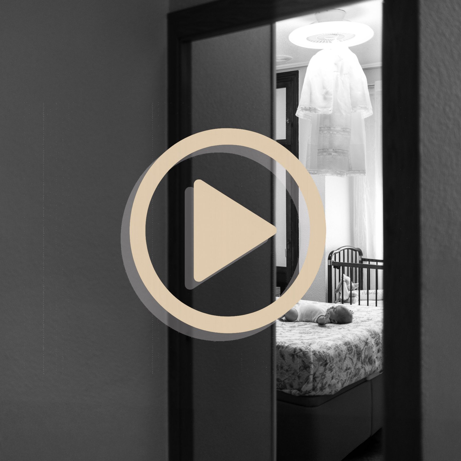 Imagen de bebé en habitación con traje de bautizo colgado, fotografía en blanco y negro con icono de reproducción de vídeo
